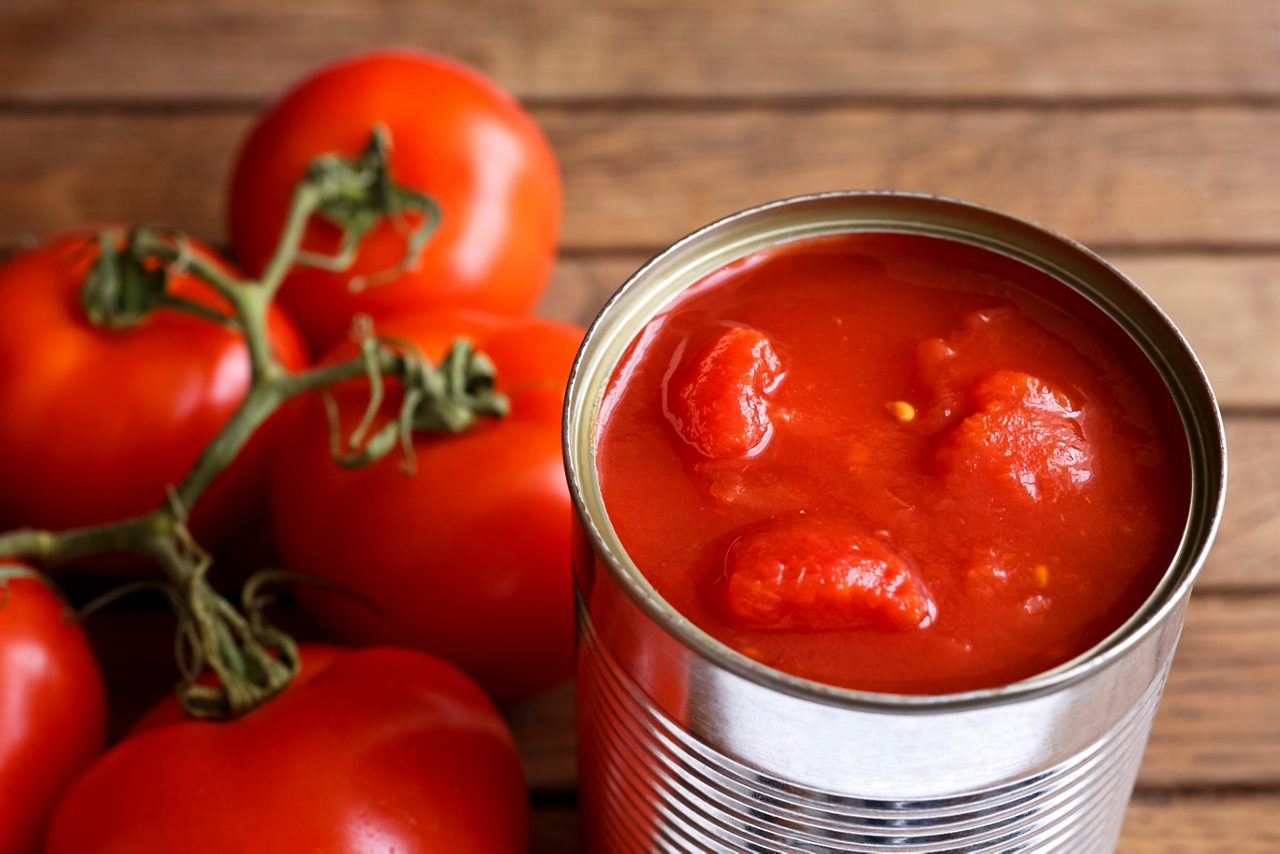 Open tin of chopped tomatoes with whole fresh unfocused tomatoes behind. Wood surface.