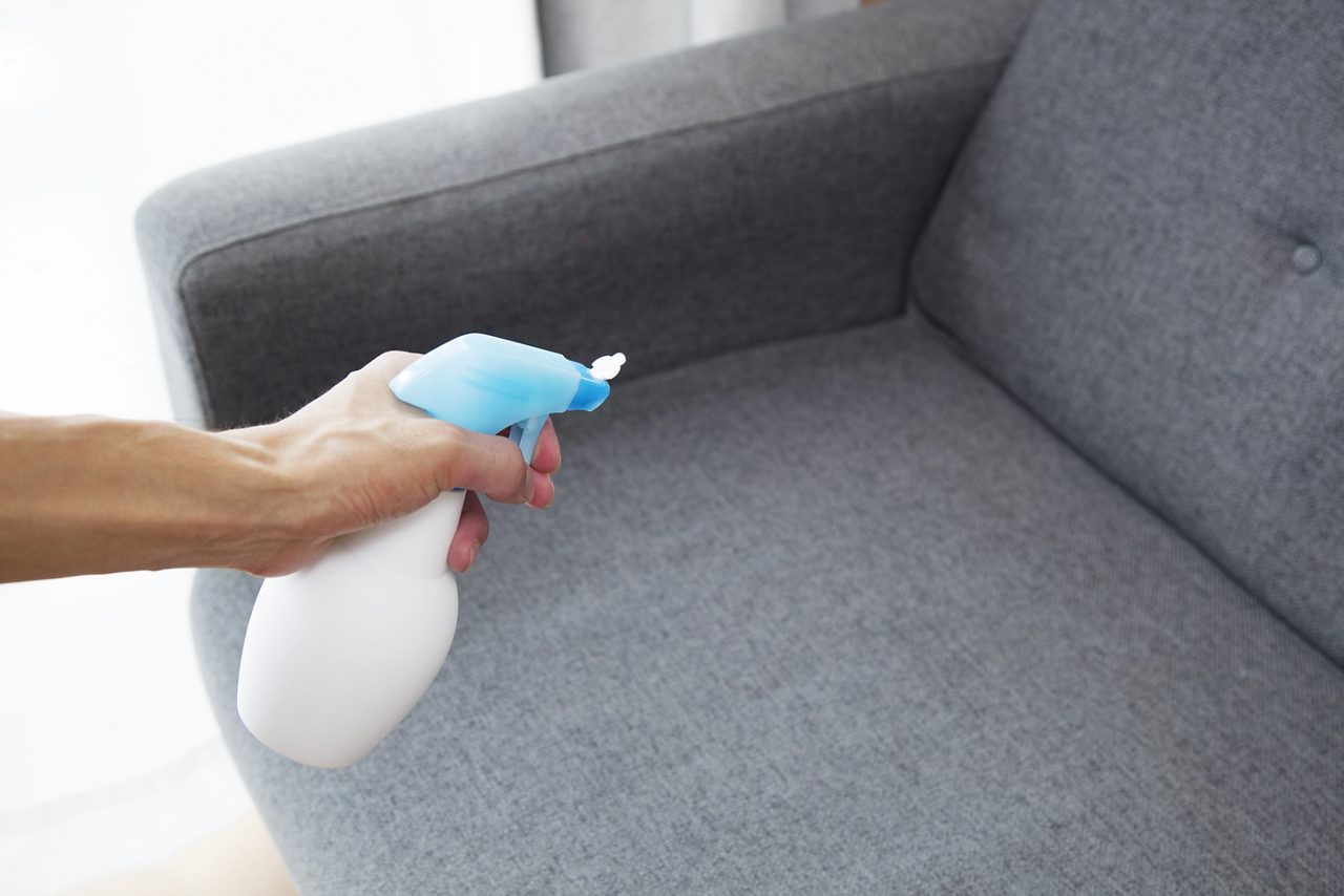 Man's hand spraying gray sofa or couch with white and blue bottle of fabric freshener or cleaning sanitizer