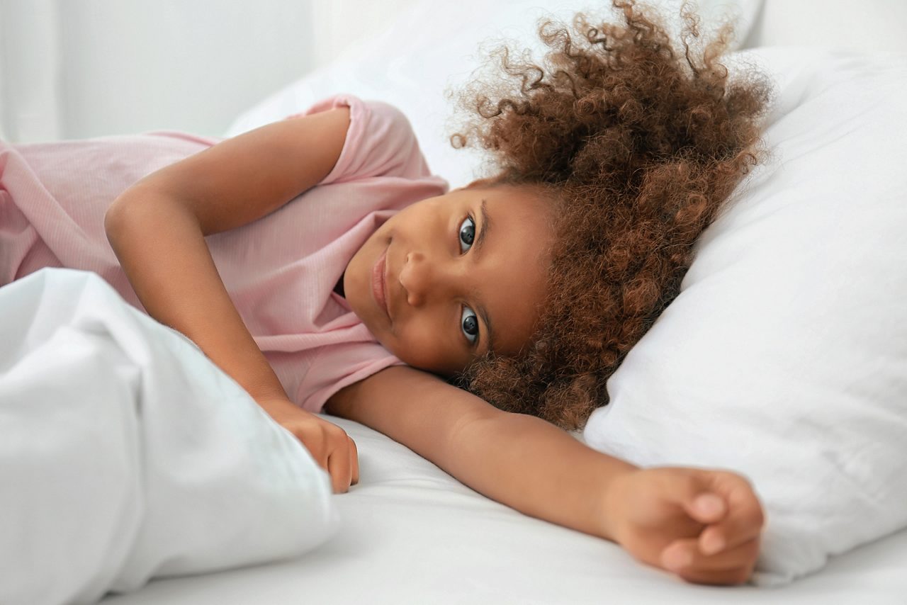 Portrait of little African-American girl smiling at camera while lying in bed with white linens