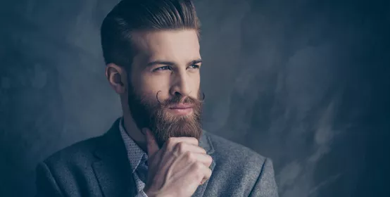 Portrait of handsome stylish young man with mustache, beard and combed back hairstyle keeps calm and thinks while holding beard and chin with hands.