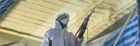 Technician spraying foam insulation using Plural Component Gun for polyurethane foam - Repair tool in the white protect suit applies a construction foam from the gun to the roof of a warehouse.