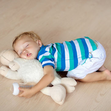 Baby sleeping on wooden floor with stuffed toy sheep and milk bottle.