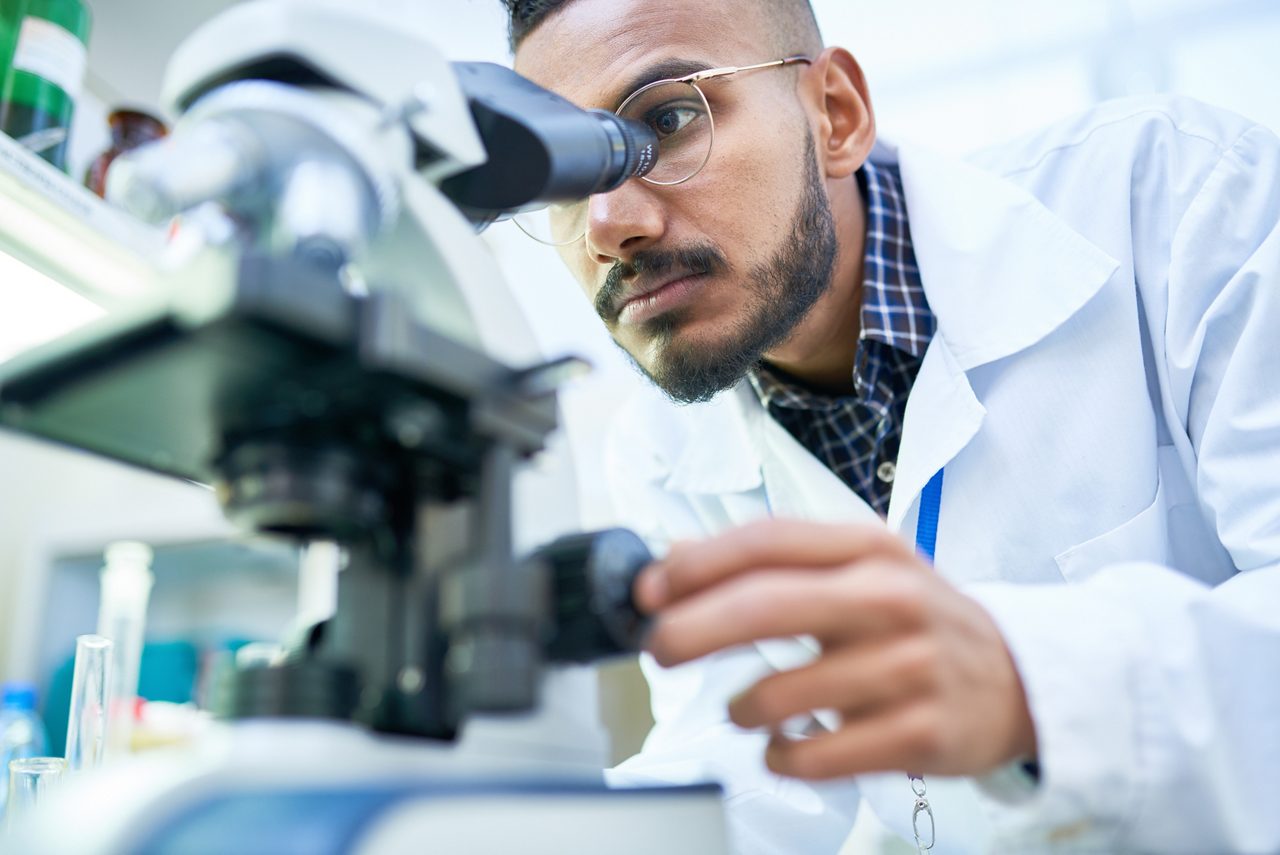 Portrait of young Middle-Eastern scientist looking in microscope while working on medical research in science laboratory, copy space.