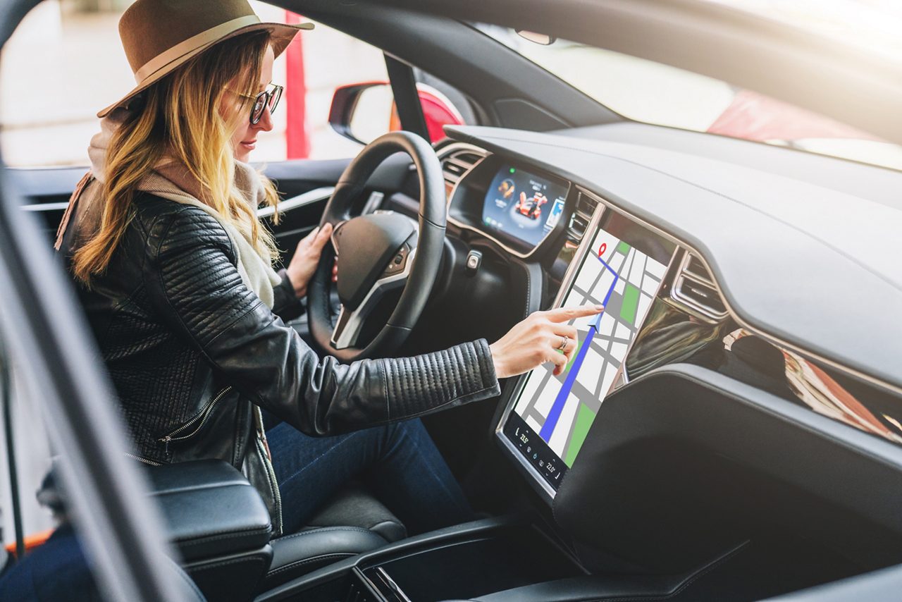 Young woman sits behind wheel in car and uses an electronic dashboard, tablet computer