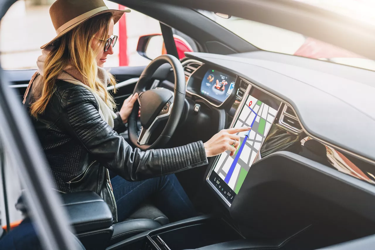 Young woman sits behind wheel in car and uses an electronic dashboard, tablet computer. Girl is traveler looking for a way through navigation system. Trip, caravanning, tourism, journey.