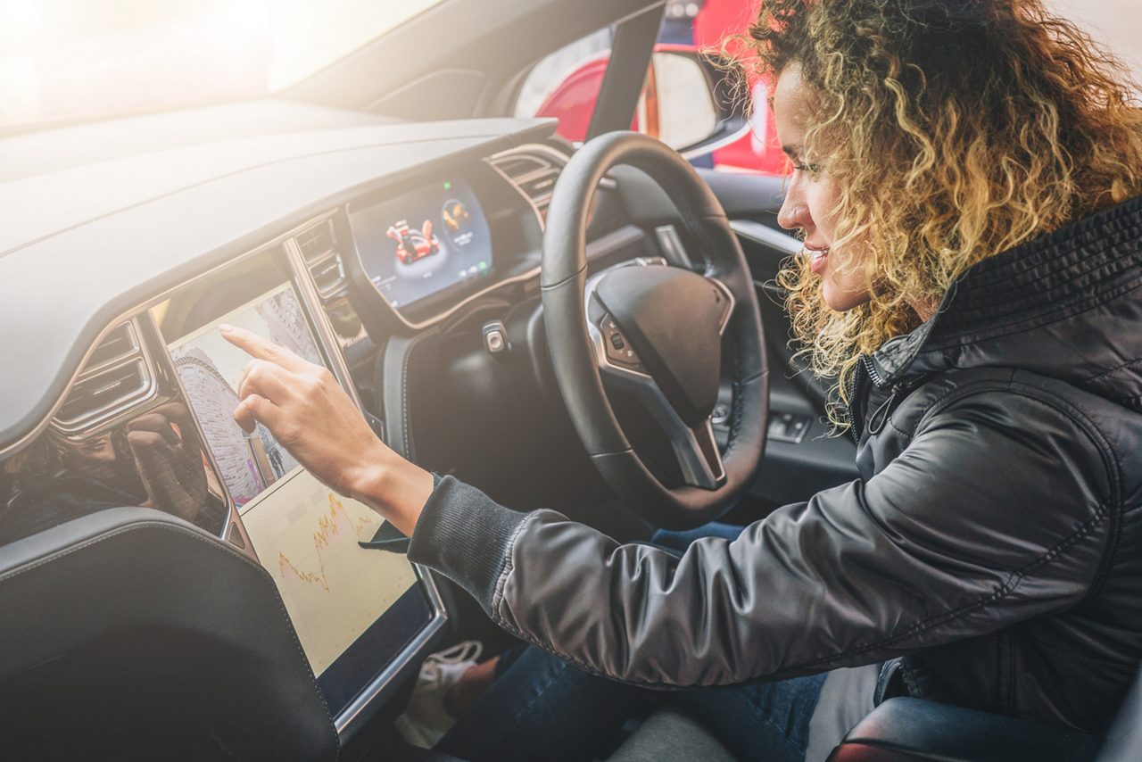 Young woman sits behind wheel in car and uses an electronic dashboard, tablet computer