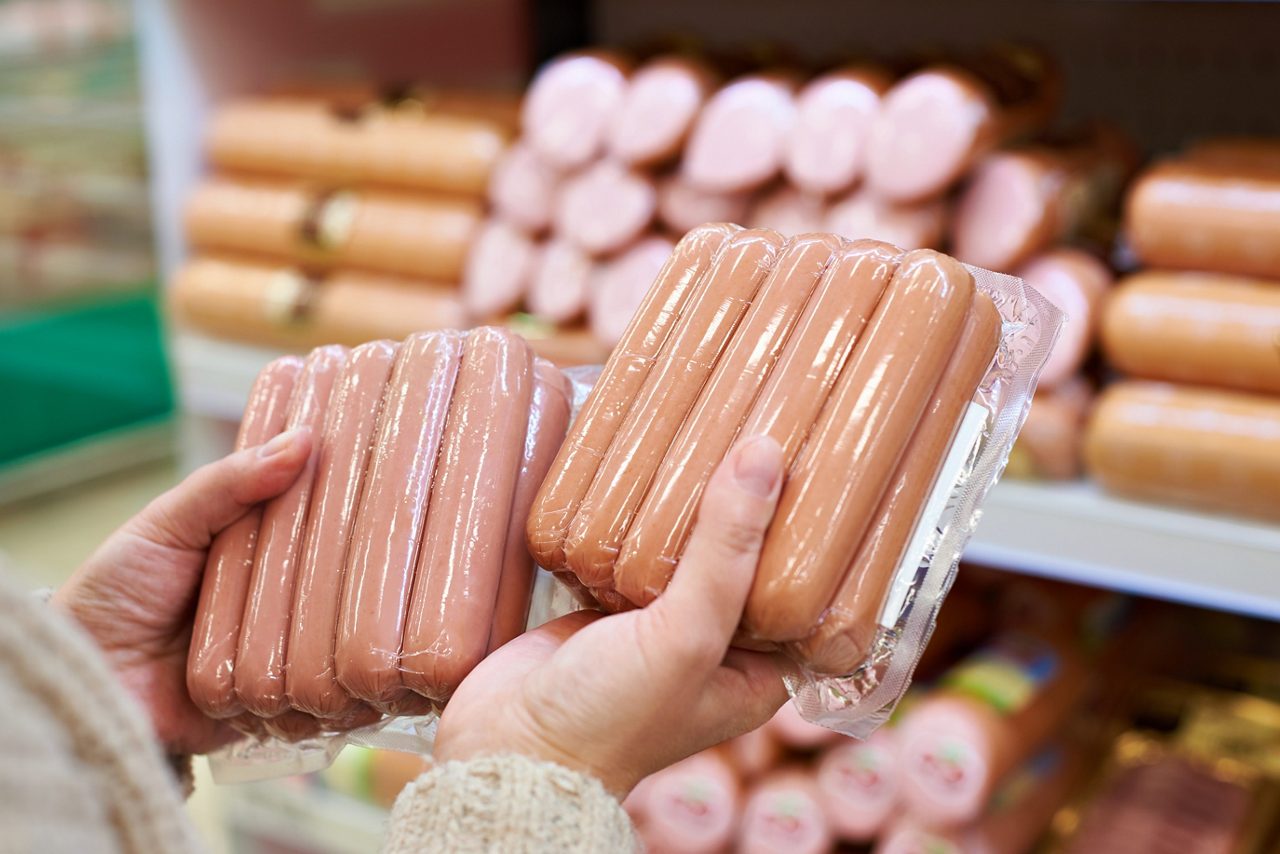 Close up of pair of hands holding two different shrink wrapped vacuum sealed packages of hot dogs with grocery store meat wall in background