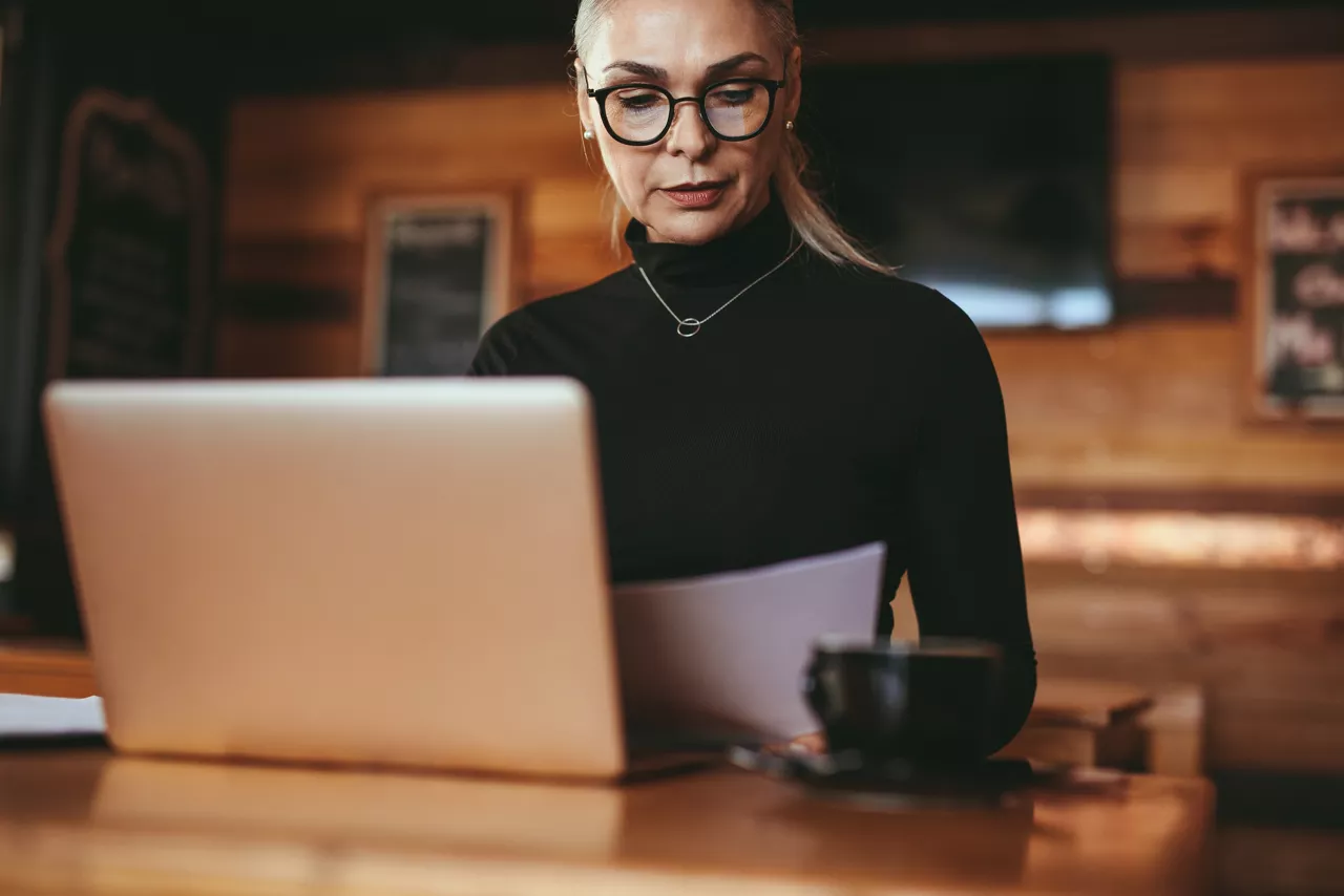 Senior businesswoman reading some documents at coffee shop. Female in casuals sitting at cafe table with laptop reading business report.