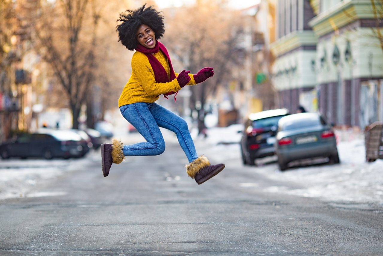 Cheerful young black woman in scarf and sweater is jumping in the middle of the street.