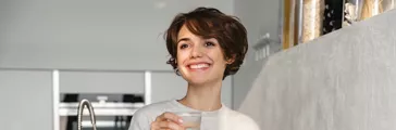 Smiling young woman holding glass of water while standing at the kitchen