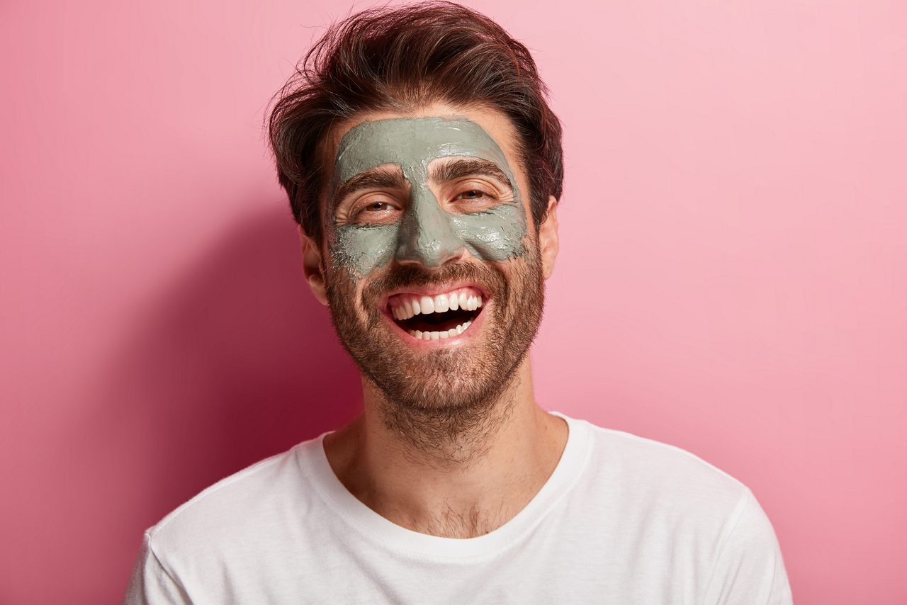 Joyful delighted man has clay mask on face, enjoys spa treatments, has broad smile, being in high spirit, cares about beauty, wears white t shirt, isolated on pink wall. Skin care and wellness concept