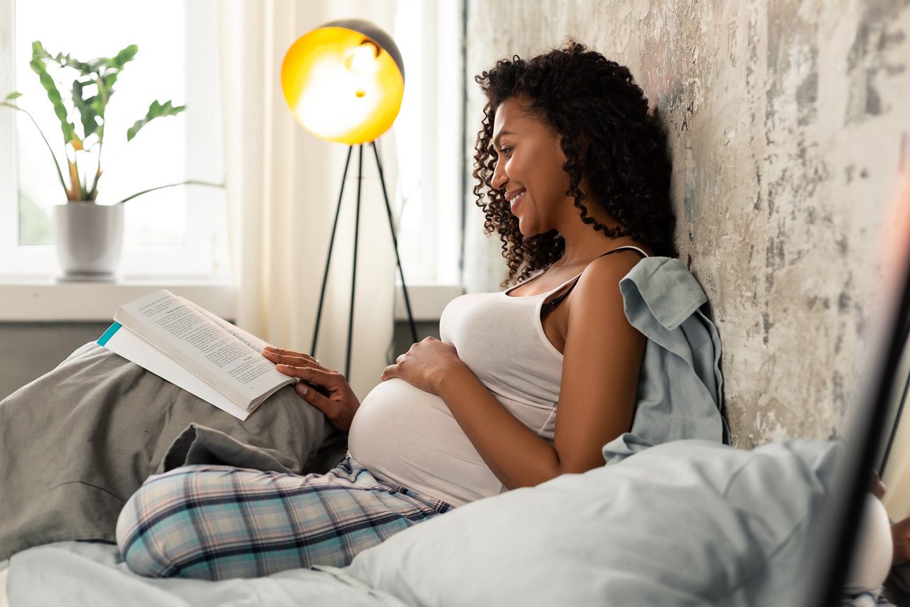 Alegre mujer embarazada sentada en su cama apoyada sobre su almohada y enrojeciendo un libro.