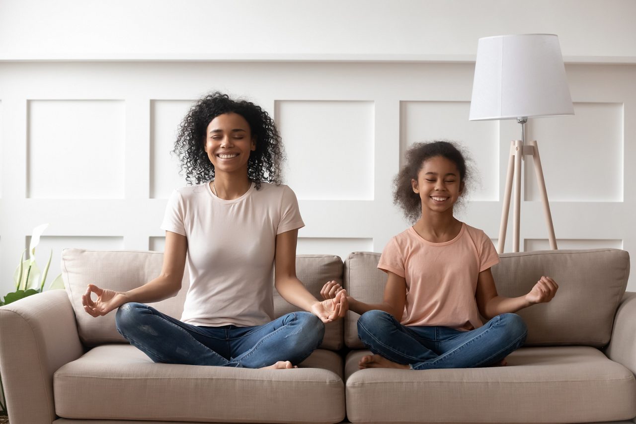 Smiling calm african family healthy mom teaching yoga cute little kid daughter doing exercises sit on sofa in lotus pose, happy mindful mixed race mother and child girl meditating together at home