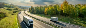 Trucks driving on the asphalt highway between deciduous forest in autumn colors under the radiant sun and dramatic clouds. View from above.