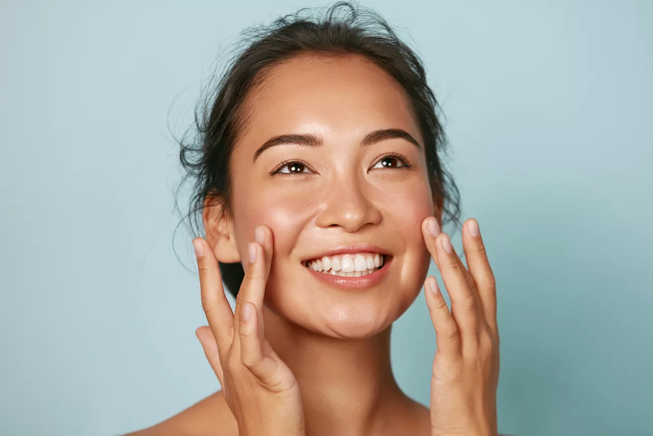 Beauty face. Smiling asian woman touching healthy skin portrait. Beautiful happy girl model with fresh glowing hydrated facial skin and natural makeup on blue background at studio. Skin care concept 