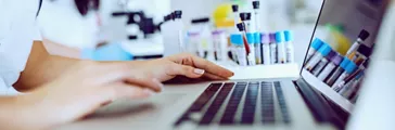 Close up of female lab assistant in white uniform sitting in lab and using laptop 