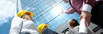 Below view of business partners in helmets looking at modern office building against cloudy sky