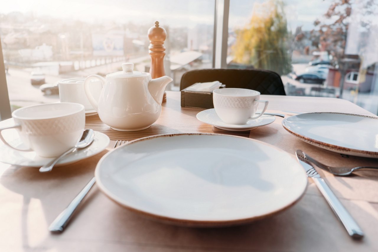 Table setting for breakfast, lunch in sun light. Dinner plate setting with empty plates, cups, teapot near window in cafe, restaurant.
