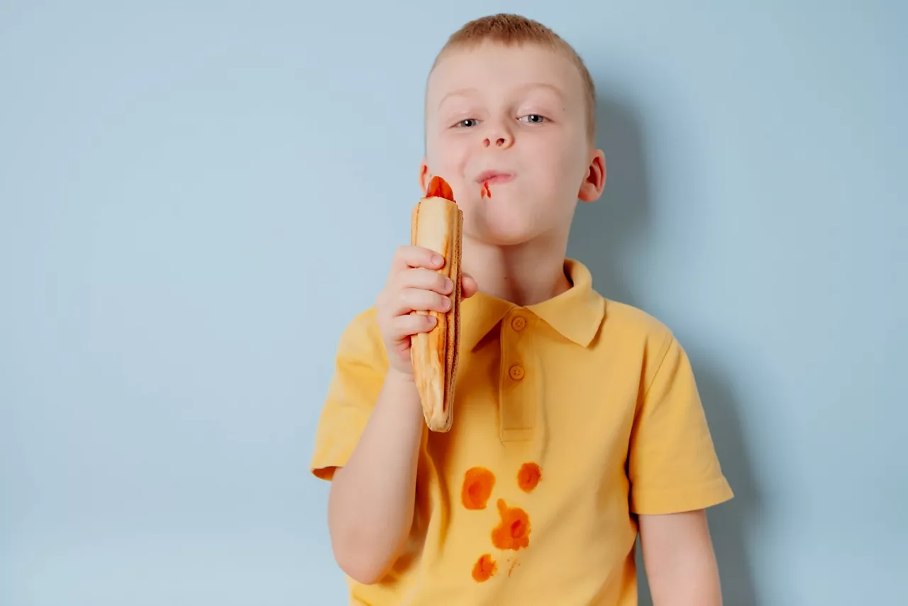 Little boy eating dog with ketchup stain on shirt  