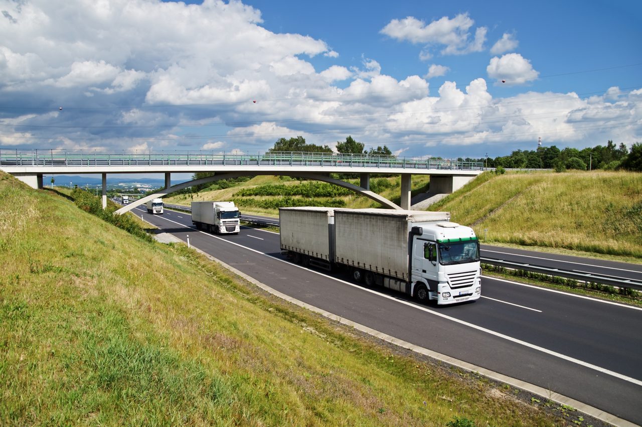 Semi trucks driving on the highway go under a concrete bridge in the countryside. View from above. Sunny day with blue sky and white clouds.