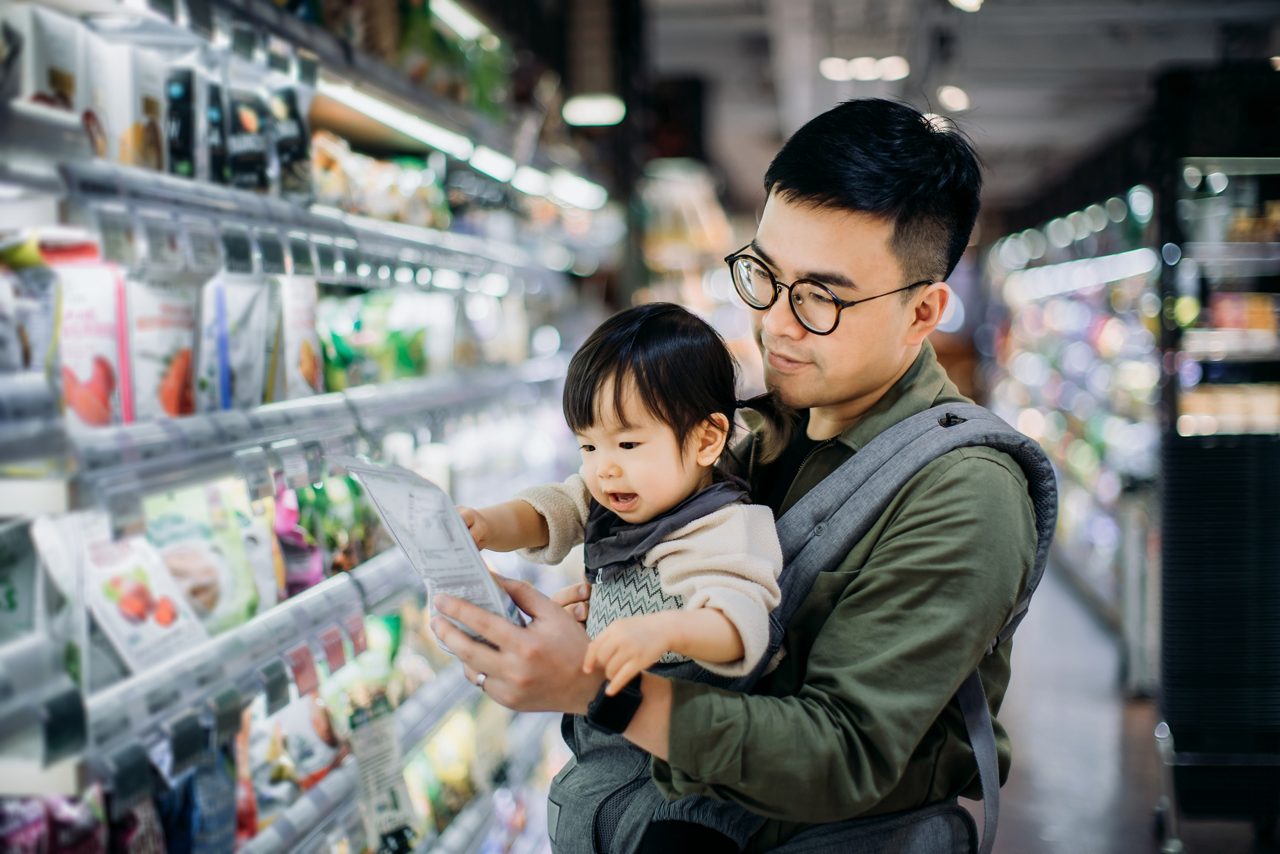 Hombre sosteniendo a un bebé mirando un producto refrigerado en el pasillo del supermercado 