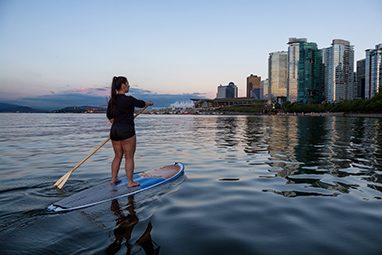 Mulher de pé em uma prancha de paddleboard olhando para os arranha-céus da cidade no horizonte.