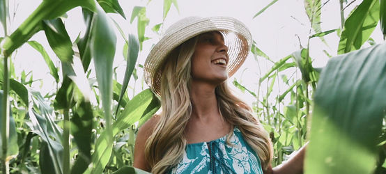 Joven mujer rubia con vestido y sombrero caminando por un campo de maíz.