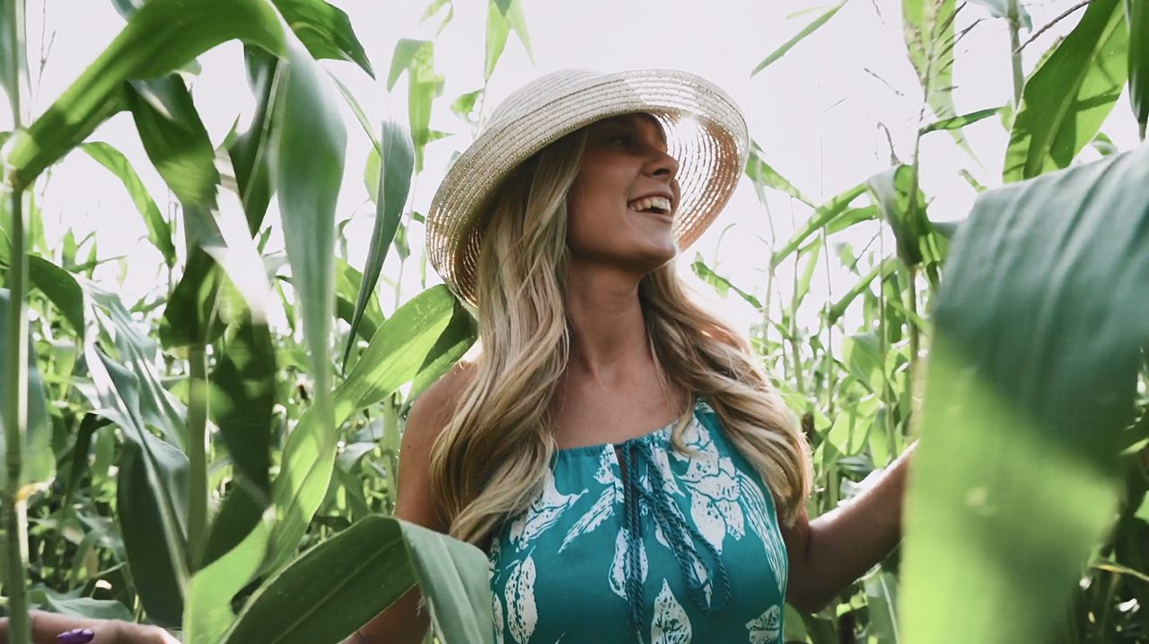 Young blonde woman wearing dress and hat walking through a corn field.