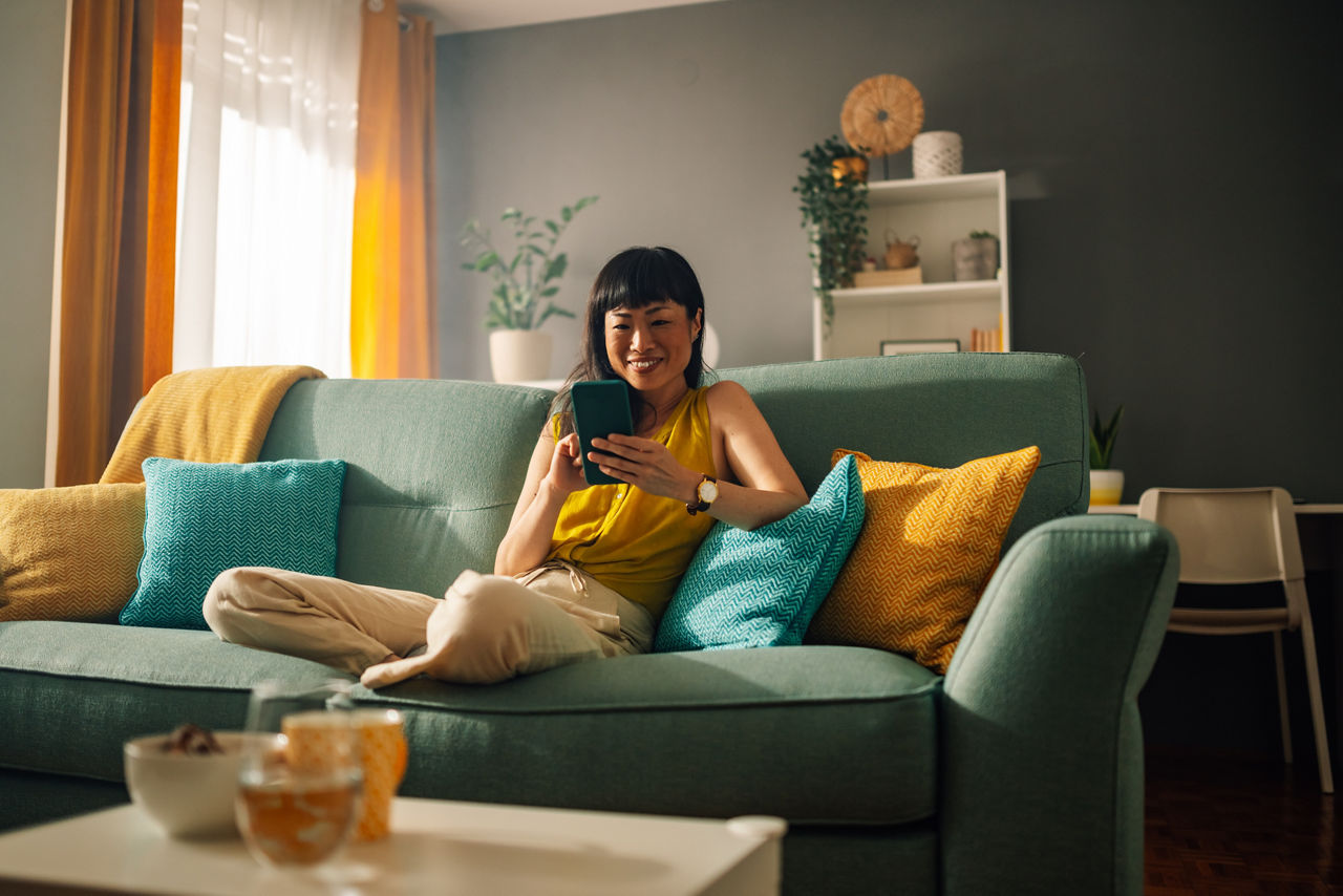 A joyful asian woman uses her smartphone while lounging on a green couch in a warm and stylish room. The room features bright yellow and green pillows, various plants, and shelves filled with decor.