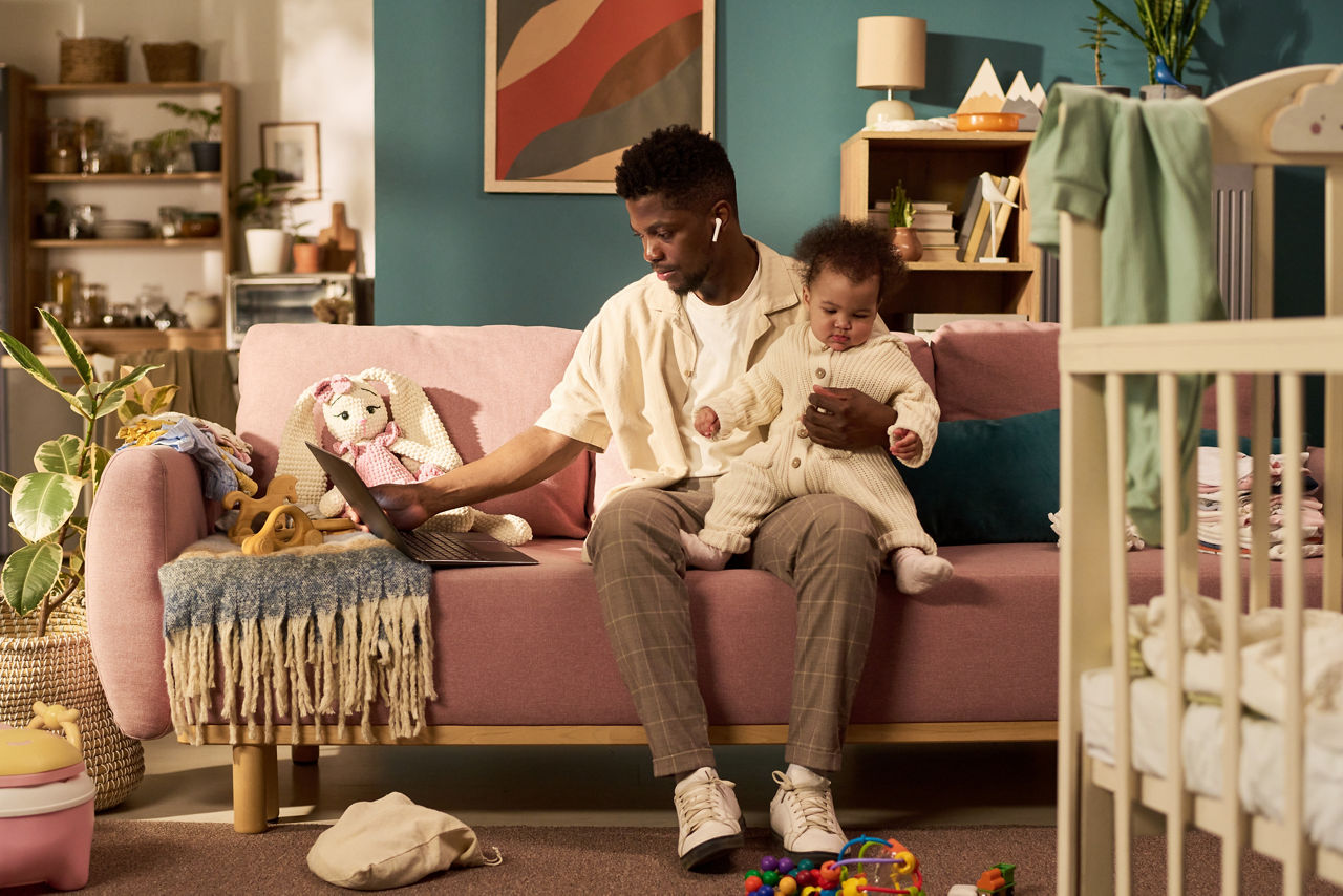Black young adult man sitting on sofa holding infant child, while working on laptop multitasking during paternity leave in home setting with toys and crib visible in background