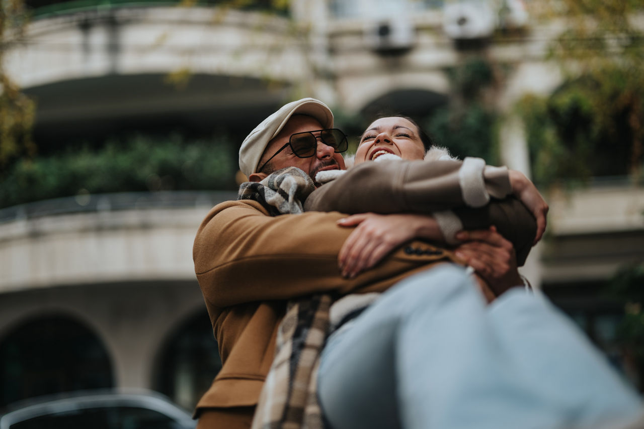 A happy couple enjoys a warm embrace in an outdoor urban environment, radiating joy and affection amidst the autumn scenery.