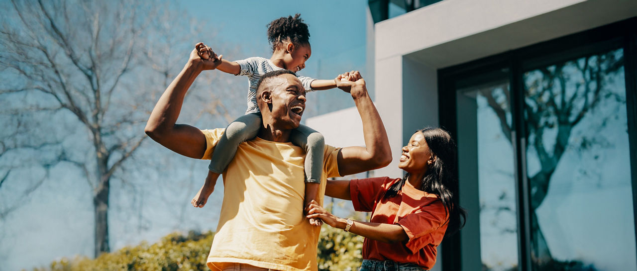 Smiling parents with their young daughter on the fathers shoulders outside their home. Highlights parenthood, love, and the joy of family connection.
