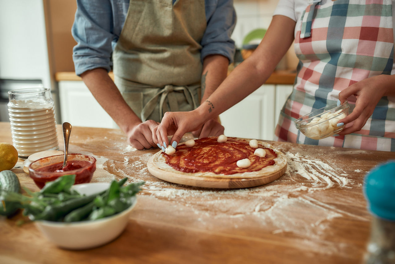 For cheesy taste. Cropped shot of couple making pizza together at home. Man in apron adding, applying tomato sauce on the dough while woman adding mozzarella cheese. Selective focus. Horizontal shot