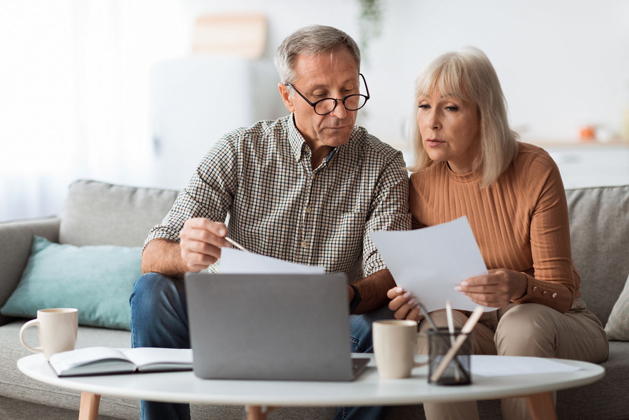 Senior Couple Looking Through Papers Using Laptop Reading Bills And Counting Expenses Together Sitting On Couch At Home. Household And Finances, Retirement Lifestyle Concept