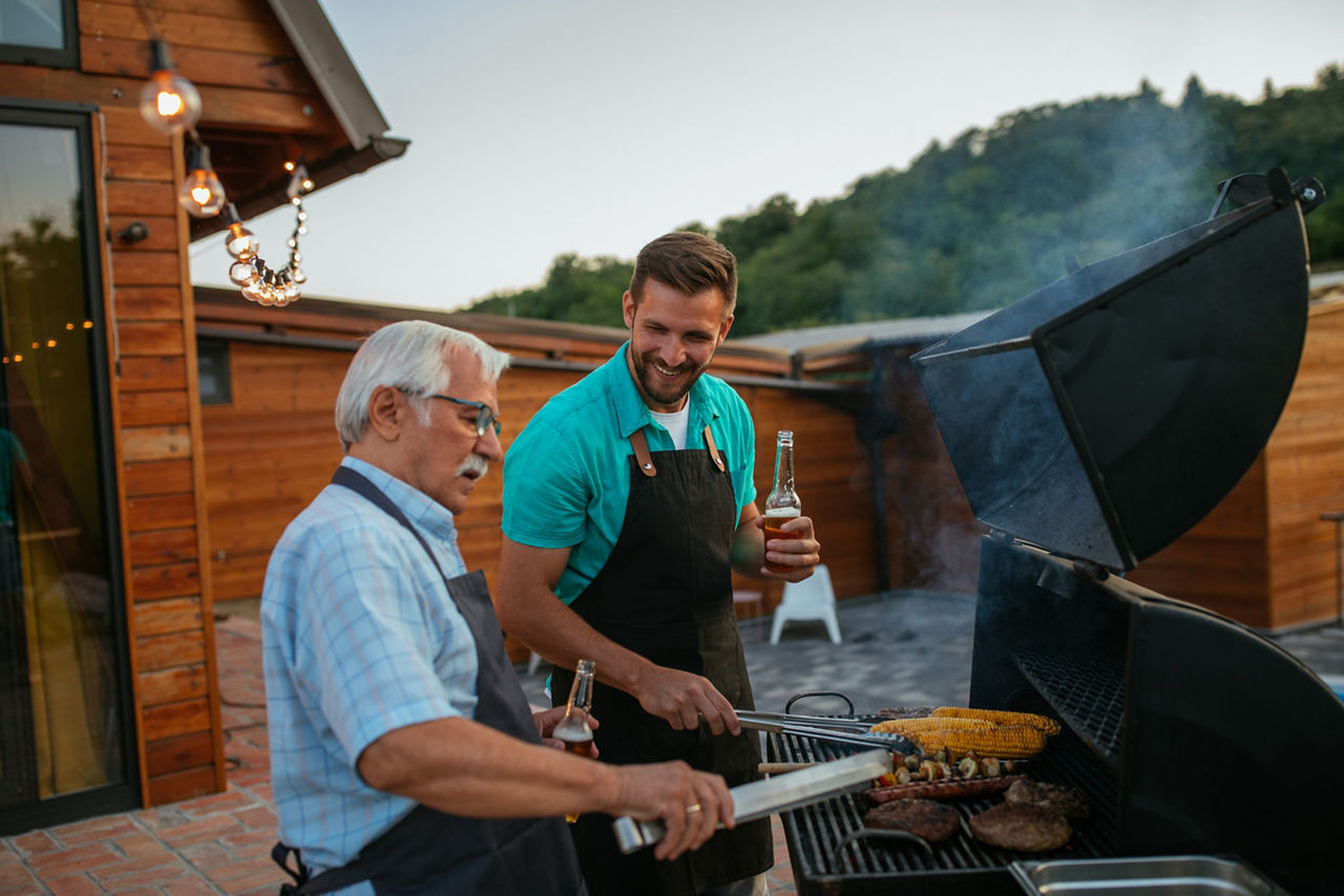 Pensioner and his son wearing aprons, holding bottles of beer and roasting meat on barbecue in the backyard