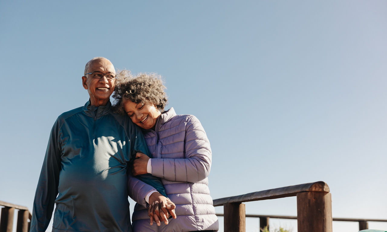 Cheerful senior couple smiling and holding hands while taking a walk along a wooden foot bridge. Romantic elderly couple enjoying a relaxing seaside holiday after retirement.