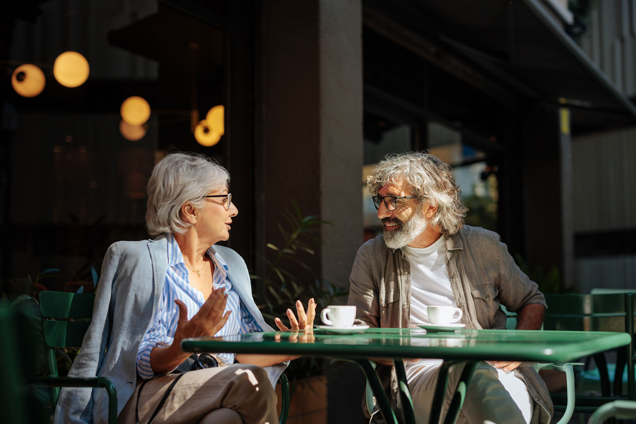 A stylish senior couple is outside sitting in a cafe having a cup of coffee and talking together.