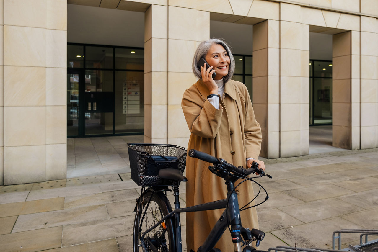 Grey-haired asian woman smiling and using cellphone while standing with bicycle outdoors