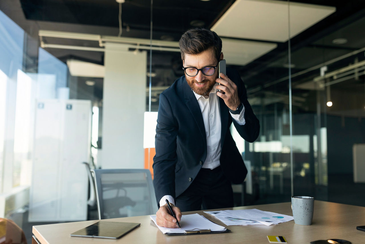 Corporate communication. Happy male manager working with papers and talking on cellphone, taking notes at financial documents, standing near workplace in office, free space