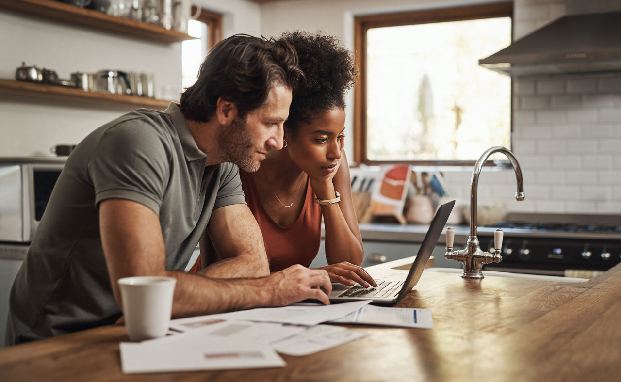 Laptop, couple and financial planning in a kitchen with documents for budget, savings and paying bills. Interracial, online and people with paperwork for taxes, mortgage and home loan application.