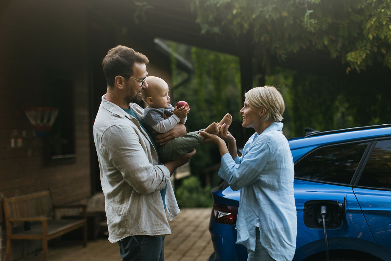 Young family with little baby standing by their electric car. Electric vehicle with charger in charging port.