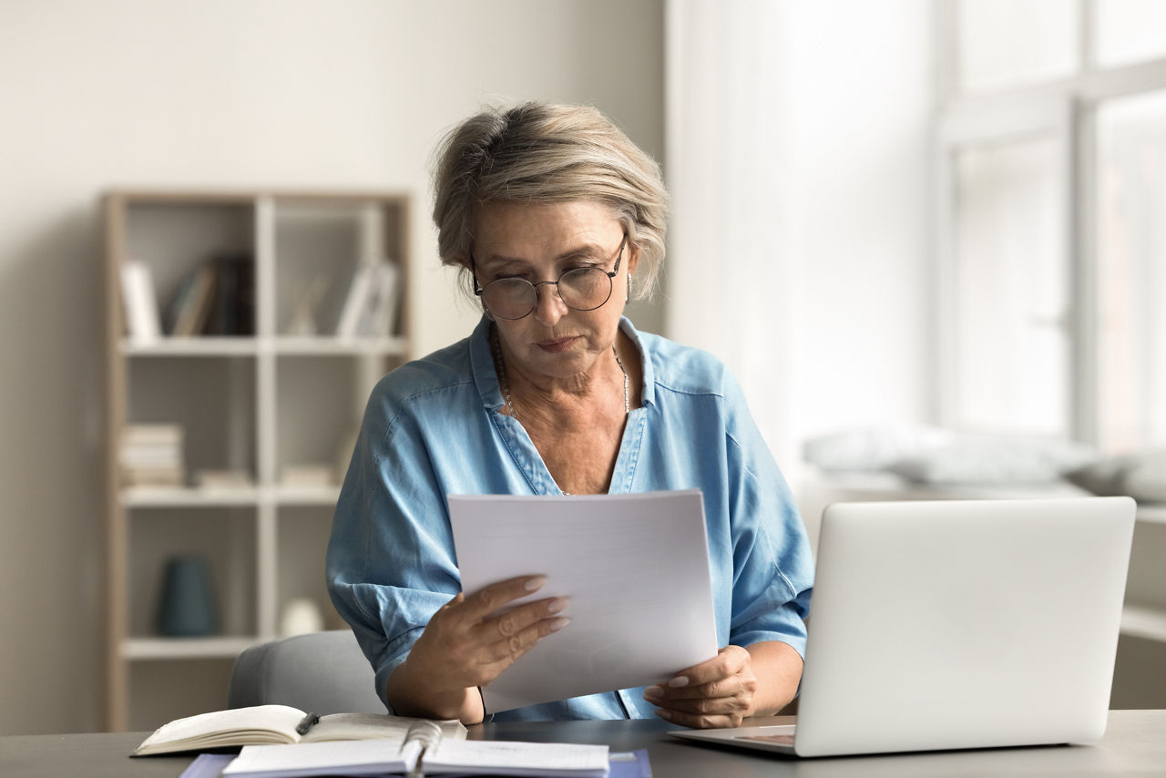 Older woman with serious expression read document sitting at desk with laptop, dealing with important paperwork, managing finances or health-related issues, learn written information looks concerned