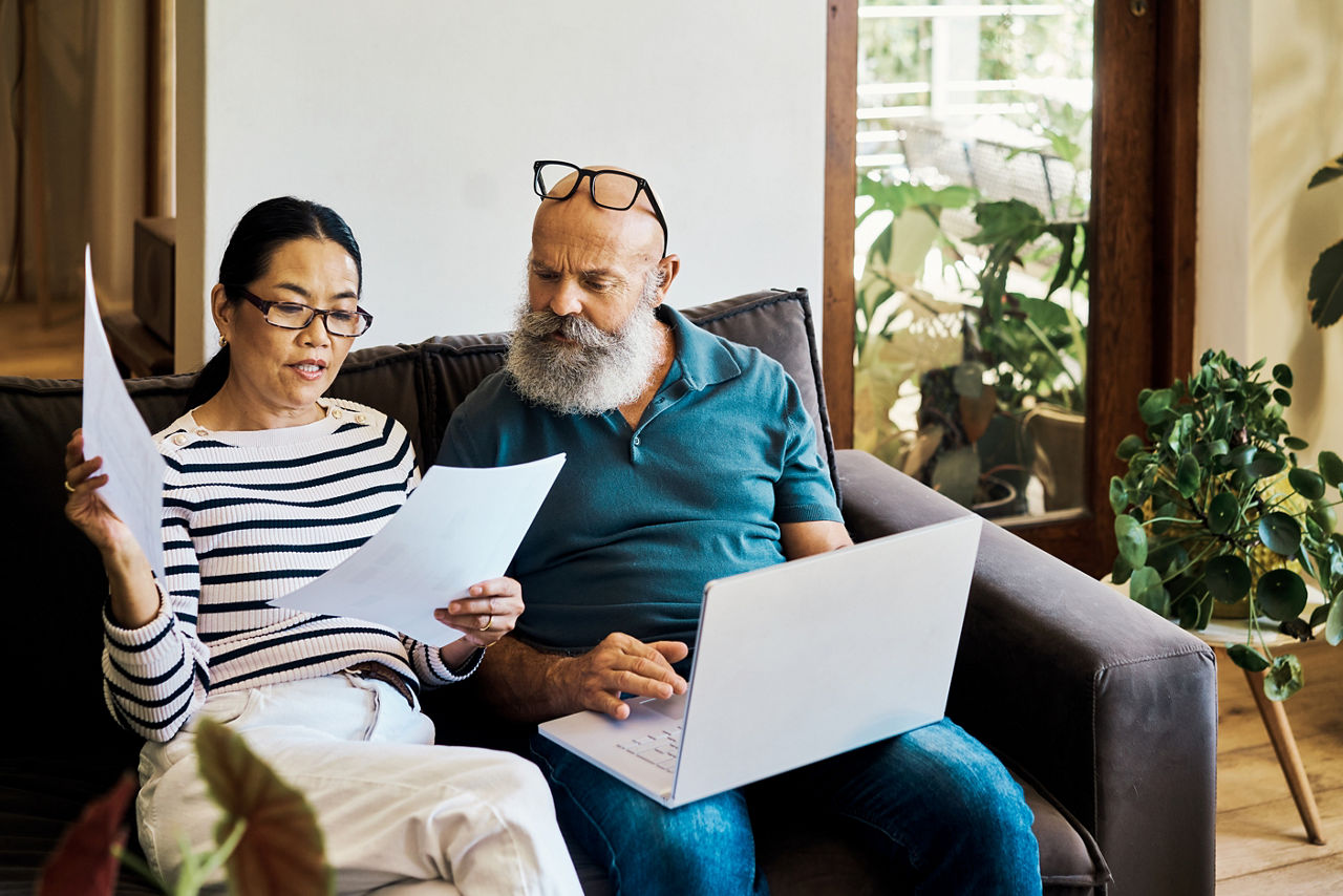 Senior interracial couple using a laptop and going retirement documents online on the couch at home. Mature husband and wife talking and reading paperwork while using a pc on the sofa in the lounge