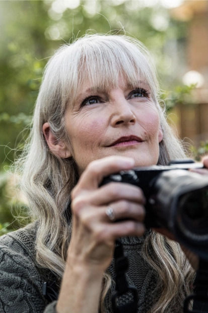 An older woman stands outside holding a camera as she looks into the distance.