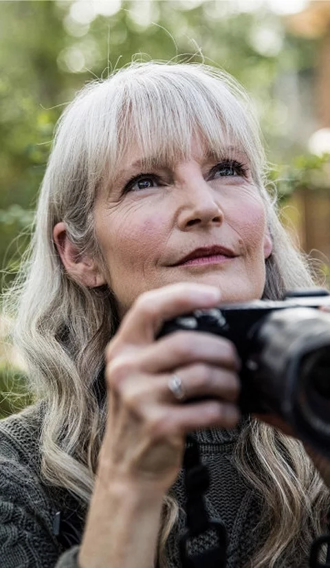 An older woman stands outside holding a camera as she looks into the distance.