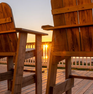 The setting sun is visible between two wooden deck chairs looking out over the ocean.