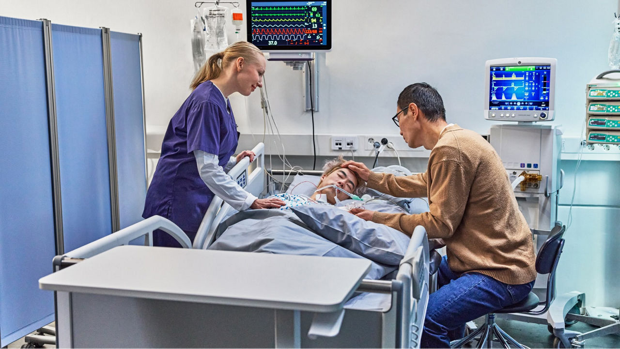 A healthcare professional and a family member stand beside a hospital bed monitoring a patient connected to GE HealthCare equipment displaying vital signs.