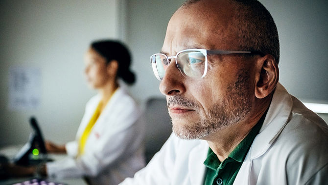 A healthcare professional focuses intently on a computer screen while another clinician works in the background.