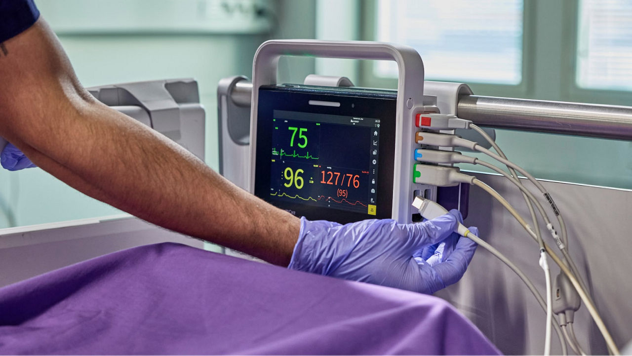 A clinician interacts with a GE HealthCare touchscreen patient monitor displaying multiple vital sign readings.