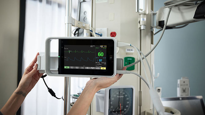 A clinician adjusts a portable patient monitoring device displaying heart rate and ECG data.